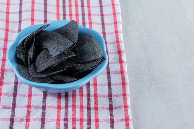 Bol bleu de chips noires croustillantes et nappe sur pierre.