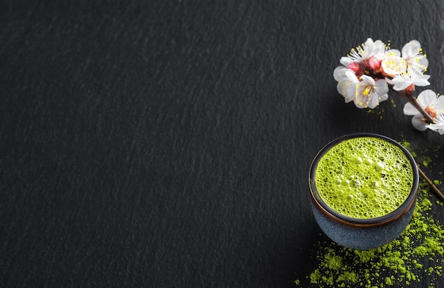 Bol de bleu avec du thé vert matcha, à côté d'une branche de cerise en fleurs et de thé en poudre sur la table