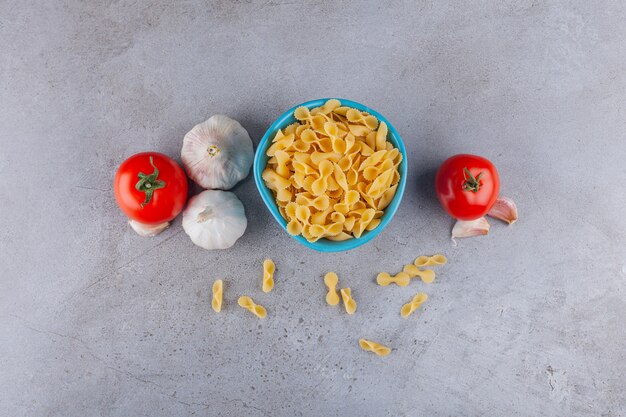 Un bol bleu plein de pâtes sèches crues Farfalle avec des légumes sur une table en pierre.