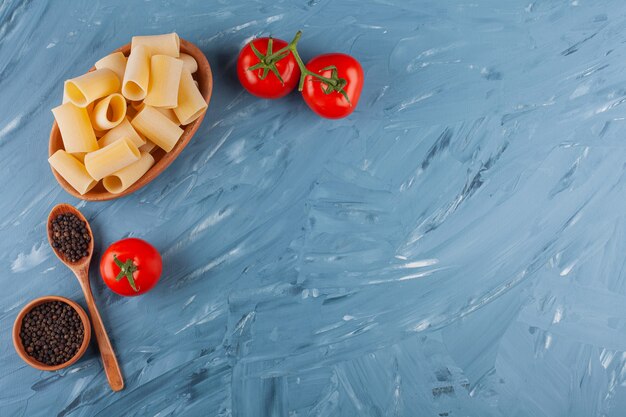 Un bol en bois de pâtes crues sèches avec des tomates rouges fraîches et des épices sur une table bleue.