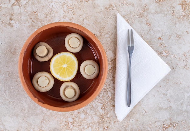 Bol de champignons et une tranche de citron à côté d'une petite fourchette sur une surface en marbre.