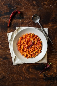 Un bol de compote de pois chiches avec de la sauce tomate chaude sur une serviette, sur fond de bois, vue aérienne. une cuillère et du piment rouge.
