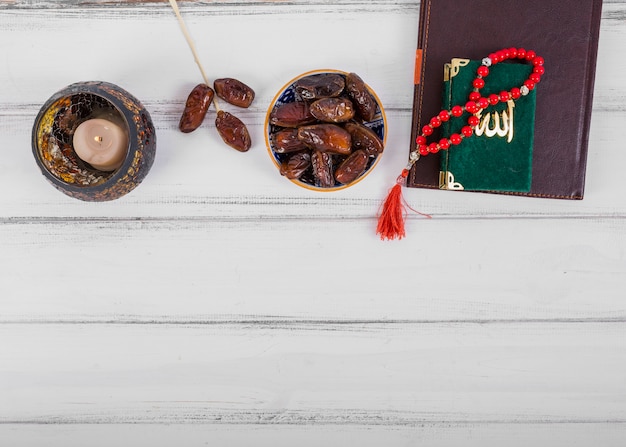 Photo gratuite bol de dattes séchées juteuses; bougie; journal intime; kuran et chapelet islamique perles de prière sur le bureau en bois blanc