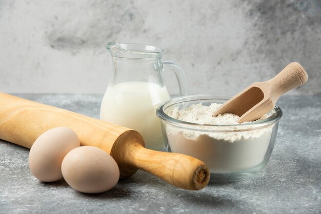 Bol de farine, œufs, lait et rouleau à pâtisserie sur table en marbre.