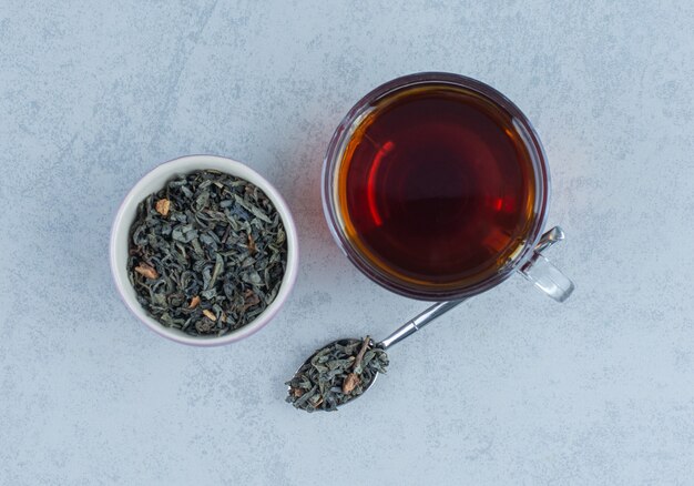 Un bol de feuilles de thé séchées et une tasse de thé avec une cuillère sur du marbre.