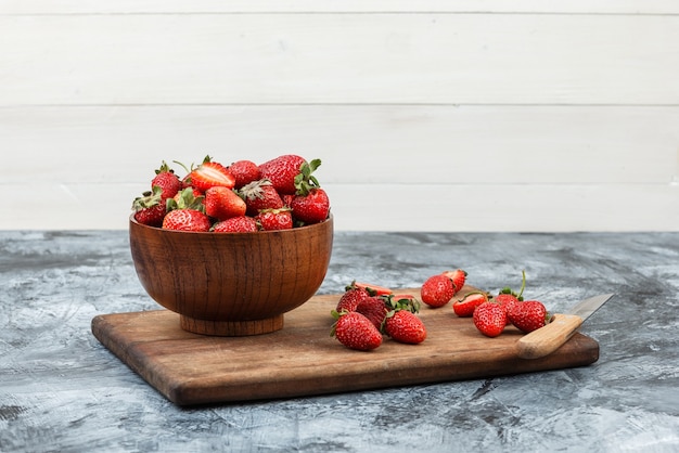 Un bol de fraises et un couteau sur une planche à découper en bois sur un fond de marbre bleu foncé et planche de bois blanc. fermer. espace libre pour votre texte