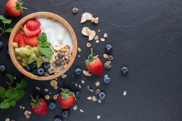 Bol de granola d'avoine avec yaourt, myrtilles fraîches, fraises, menthe kiwi et planche de noix pour un petit-déjeuner sain, vue de dessus, espace de copie, mise à plat. Concept de menu de petit-déjeuner sain. Sur le rocher noir