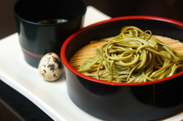 Bol noir de nouilles vertes avec oeuf de caille et une sauce servie sur un plateau blanc