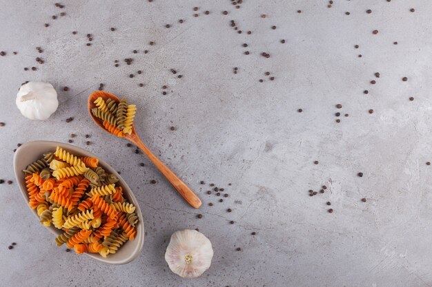 Un bol de pâtes en spirale crues multicolores avec des grains d'ail et de poivre.