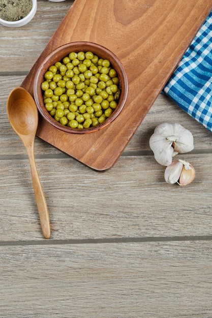 Un bol de pois verts bouillis avec une cuillère, de l'ail et une nappe bleue sur une table en bois.