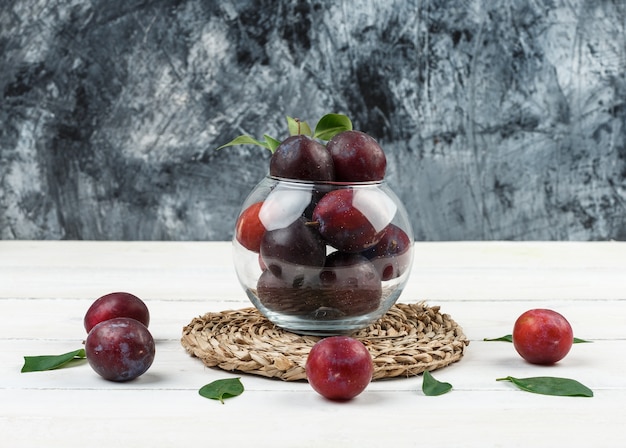 Photo gratuite un bol de prunes sur un napperon en osier sur un fond de marbre bleu foncé et planche de bois blanc. fermer.