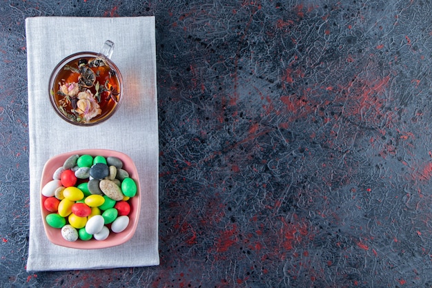 Bol rose de bonbons colorés et tasse de thé noir sur une surface sombre.