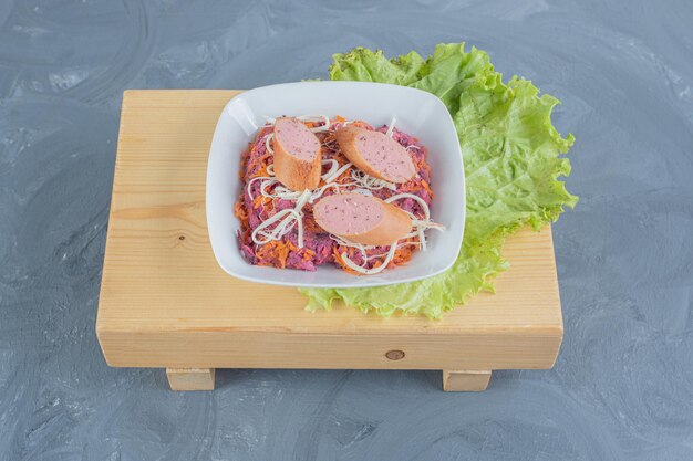 Bol de salade de noix et de betteraves sur une planche de bois avec des feuilles de laitue, garni de saucisses et de fromage sur une surface en marbre.