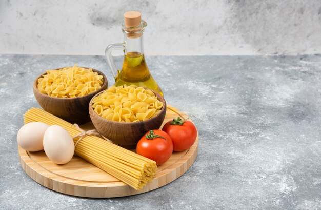 Bols de pâtes crues, œufs, tomates et huile d'olive sur une surface en marbre.