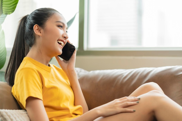 Bonheur joyeux souriant jeune adulte asiatique femme femme porter un tissu jaune conversation à la main sur smartphone sur canapé à la maison quarantaine à distance période de verrouillage fond intérieur de la maison
