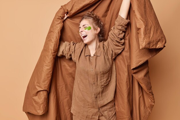 Bonjour concept Une jeune femme détendue tient des bâillements de couverture et s'étire les bras après avoir dormi applique des patchs de beauté hydrogel vêtus d'un pyjama confortable isolé sur fond marron