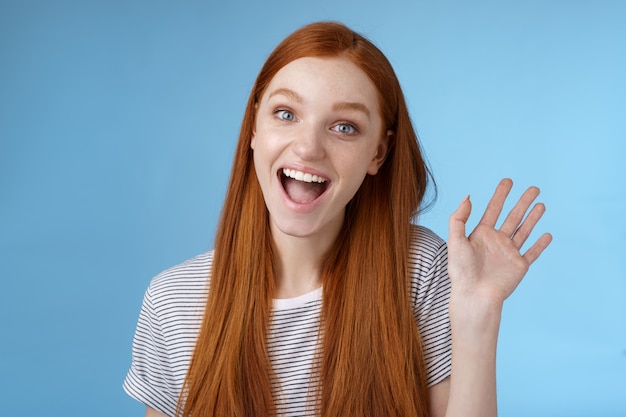 Bonjour veux être amis. Jeune femme rousse enthousiaste et mignonne faisant connaissance avec des collègues souriants heureux agitant la main levée salut geste de salutation accueillant, dites au revoir debout fond bleu.