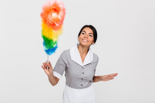 Bonne brunette femme de chambre en uniforme gris glisser avec plumeau