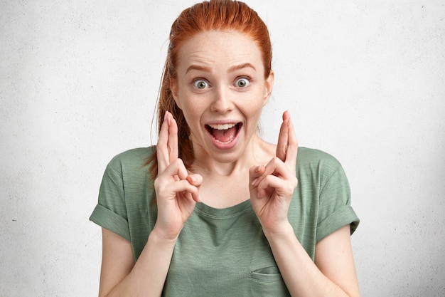 Bonne drôle de femme gingembre ravie avec noeud de cheveux, porte un t-shirt décontracté, croise les doigts avec un grand espoir, modèles en studio contre blanc