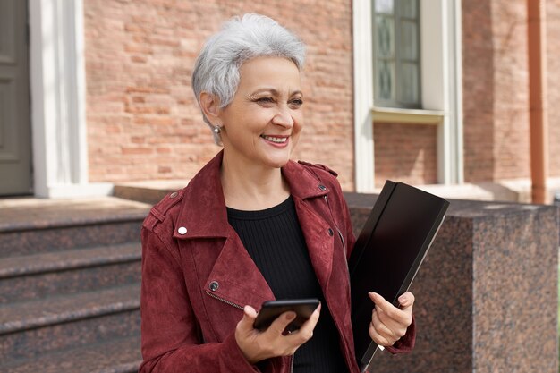 Bonne femme de 50 ans réussie en veste tendance posant sur la porte tenant un téléphone intelligent et un dossier