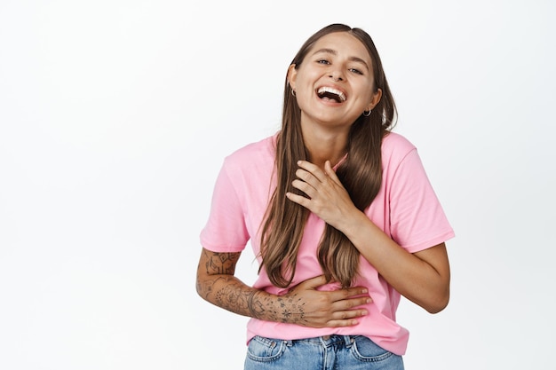 Bonne fille candide riant, souriant et riant, regardant quelque chose de drôle, debout avec les mains sur la poitrine, portant un t-shirt rose, fond blanc.