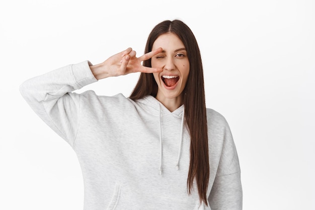 Bonne fille kawaii montrant la paix vsign et un clin d'œil en souriant et en regardant la caméra positive debout en sweat-shirt sur fond blanc Espace de copie