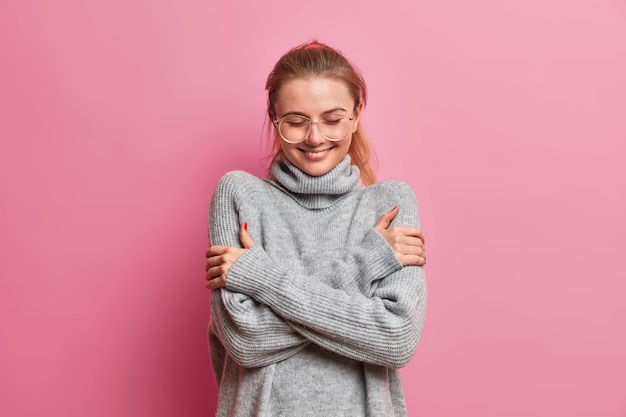 Bonne fille tendre vêtue d'un pull surdimensionné chaud gris, s'embrasse, apprécie le confort pendant la journée pluvieuse d'automne, porte des lunettes transparentes sourit agréablement