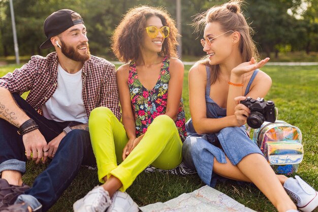 Bonne jeune entreprise d'amis assis dans le parc, l'homme et la femme s'amusant ensemble, style de mode hipster d'été coloré, voyageant avec appareil photo, parlant, souriant