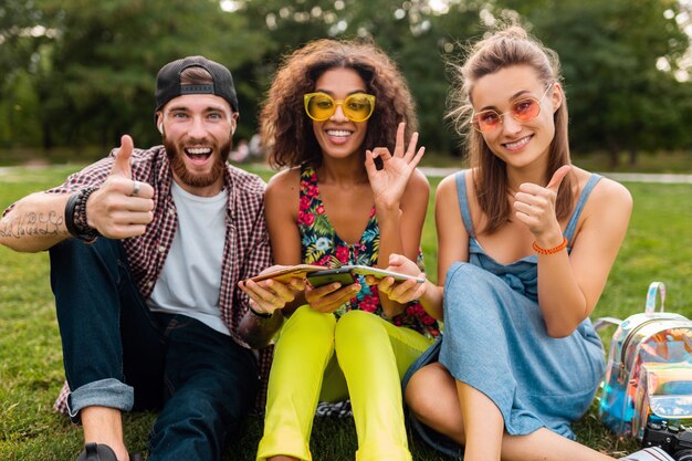 Bonne jeune entreprise d'amis souriants assis dans le parc à l'aide de smartphones, s'amuser ensemble