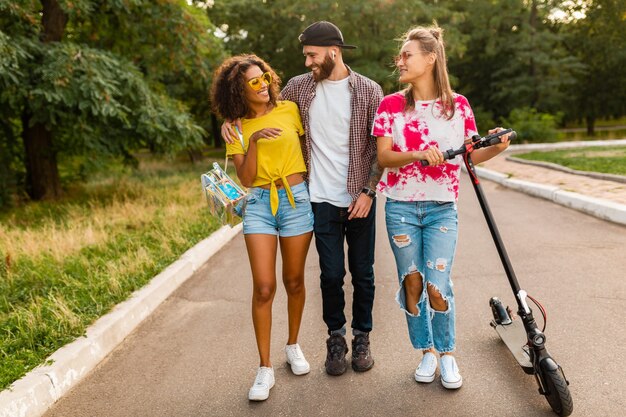 Bonne jeune entreprise d'amis souriants marchant dans le parc avec scooter électrique, homme et femme s'amusant ensemble