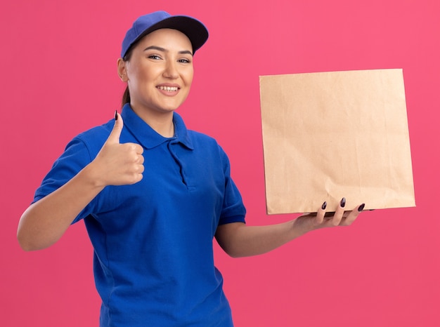 Bonne jeune femme de livraison en uniforme bleu et cap tenant le paquet de papier à l'avant souriant joyeusement montrant les pouces vers le haut debout sur le mur rose