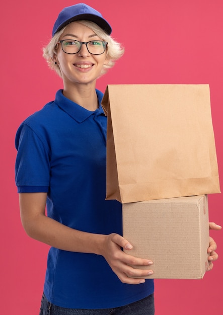 Bonne jeune femme de livraison en uniforme bleu et cap tenant le paquet de papier et boîte en carton souriant joyeusement sur mur rose