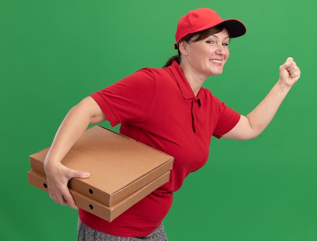 Bonne jeune femme de livraison en uniforme rouge et cap rush en cours d'exécution pour la livraison de boîtes de pizza pour le client sur le mur vert