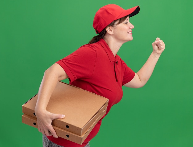 Bonne Jeune Femme De Livraison En Uniforme Rouge Et Cap Rush En Cours D'exécution Pour La Livraison De Boîtes De Pizza Pour Le Client Sur Le Mur Vert