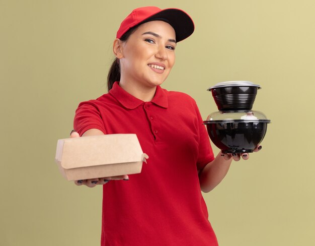 Bonne jeune femme de livraison en uniforme rouge et cap tenant des paquets de nourriture à l'avant avec le sourire sur le visage debout sur le mur vert