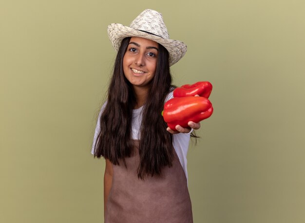 Bonne jeune fille de jardinier en tablier et chapeau d'été montrant des poivrons rouges frais avec le sourire sur le visage debout sur le mur vert