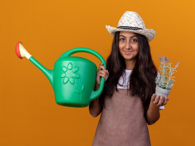 Bonne jeune fille de jardinier en tablier et chapeau d'été tenant arrosoir et plante en pot avec sourire sur le visage debout sur un mur orange