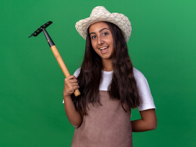 Bonne jeune fille de jardinier en tablier et chapeau d'été tenant un mini râteau avec sourire sur le visage debout sur un mur vert