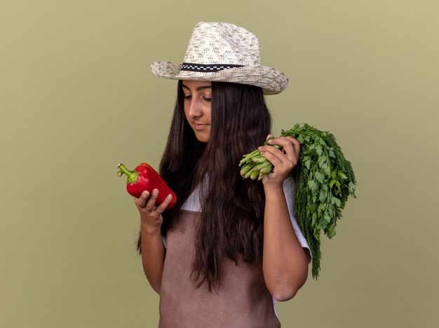 Bonne jeune fille de jardinier en tablier et chapeau d'été tenant le poivron rouge frais et les herbes fraîches à la recherche de poivre avec intérêt debout sur le mur vert