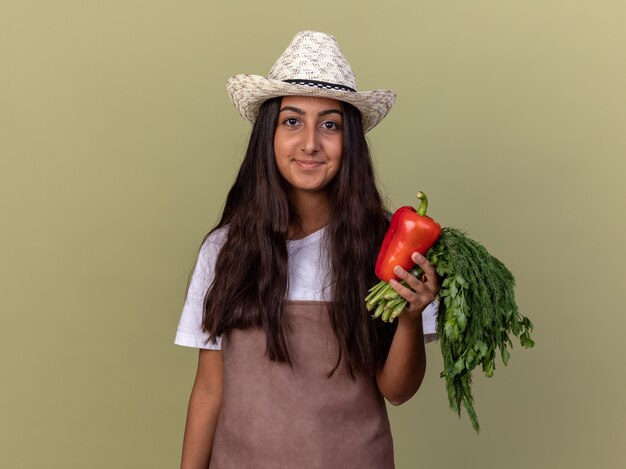 Bonne jeune fille de jardinier en tablier et chapeau d'été tenant le poivron rouge frais et les herbes fraîches souriant debout sur le mur vert