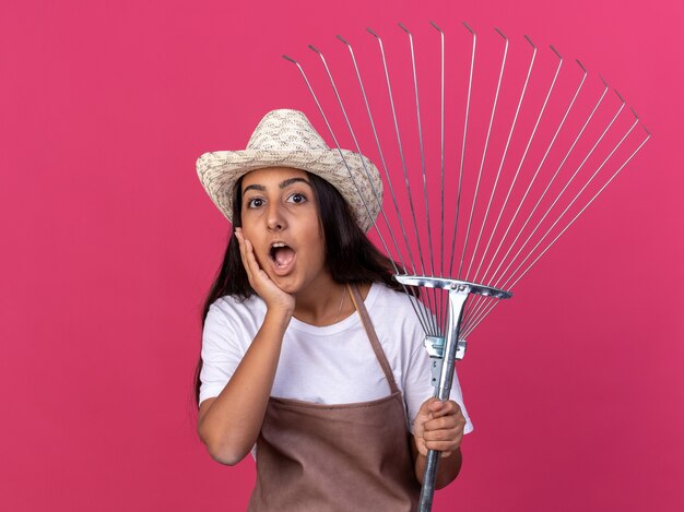 Bonne jeune fille de jardinier en tablier et chapeau d'été tenant le râteau étonné et surpris debout sur le mur rose