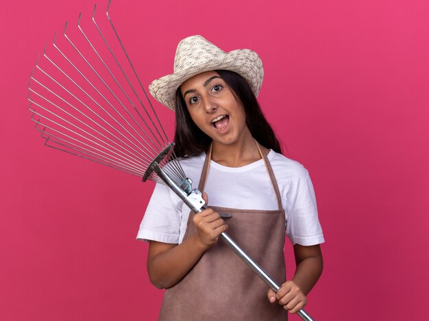 Bonne jeune fille de jardinier en tablier et chapeau d'été tenant le râteau avec le sourire sur le visage debout sur le mur rose