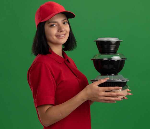 Photo gratuite bonne jeune fille de livraison en uniforme rouge et cap tenant la pile de colis alimentaires souriant sympathique debout sur le mur vert