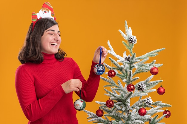 Bonne jeune fille en pull de Noël portant un bandeau drôle à côté d'un arbre de Noël sur fond orange