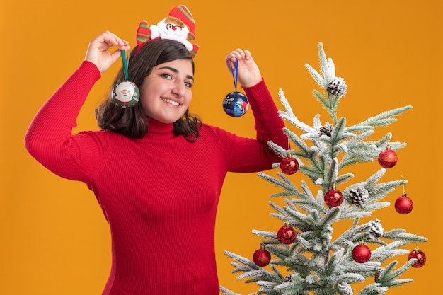 Bonne jeune fille en pull de Noël portant un bandeau drôle tenant des boules de Noël avec sourire sur le visage debout à côté d'un arbre de Noël sur un mur orange