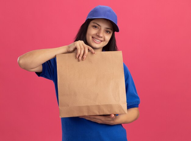 Bonne jeune livreuse en uniforme bleu et chapeau montrant le paquet de papier souriant joyeusement debout sur le mur rose