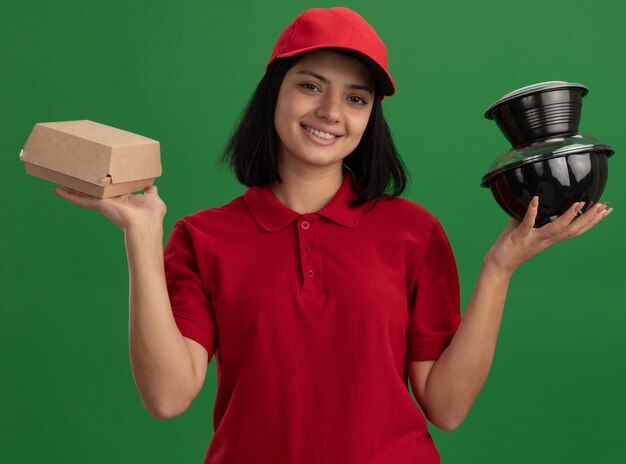Bonne jeune livreuse en uniforme rouge et cap tenant des paquets de nourriture souriant sympathique debout sur le mur vert