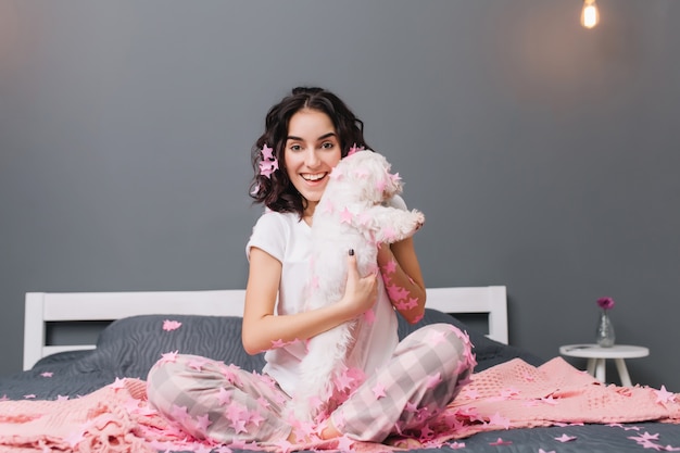 Bonne journée, vraies émotions positives de la jeune femme joyeuse en pyjama aux cheveux bouclés brune s'amusant avec petit chien en guirlandes roses sur le lit