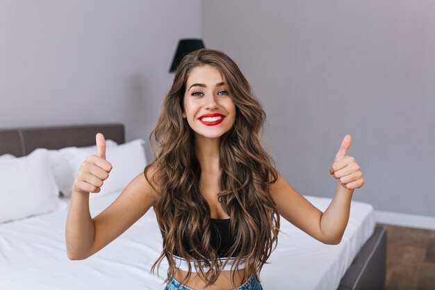 Bonne matinée de joyeuse jolie fille aux longs cheveux bruns et lèvres rouges se détendre sur le lit dans un appartement moderne. T-shirt noir, grand sourire, bonne humeur, montrant une vraie émotion, regard apprécié.