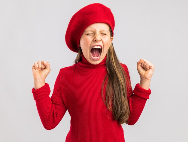 Bonne petite fille blonde portant un béret rouge gardant les poings levés les yeux fermés isolé sur mur blanc